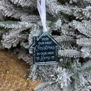 Decorative ornament shaped like a house with '2025 Our First Christmas in Our New Home' text, hanging on a Christmas tree.