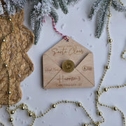 Wooden envelope-shaped ornament with 'Santa Claus' and 'Harrisons Christmas Wish List' text, surrounded by decorative elements on a white background.